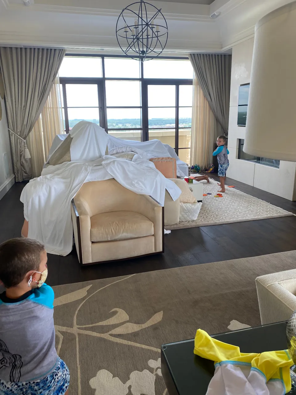 Children building a large fort from armchairs, cushions and white sheets in a living room.