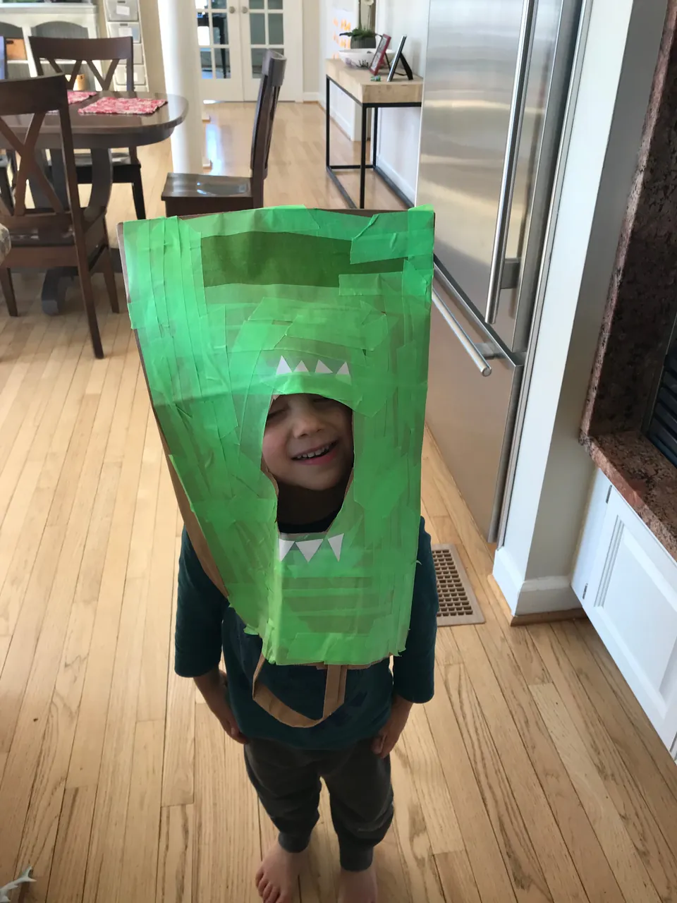 Child wearing a tall cardboard character head covered in green tape, with a cut-out face opening.