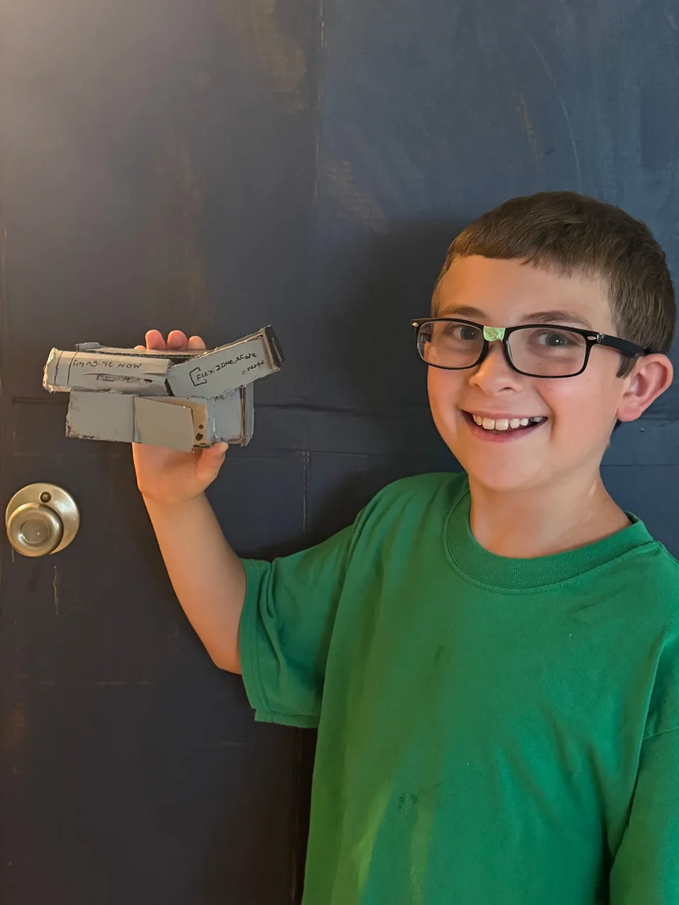 Billy, the Storyteller, in a green shirt, smiling while holding a handmade cardboard spaceship.